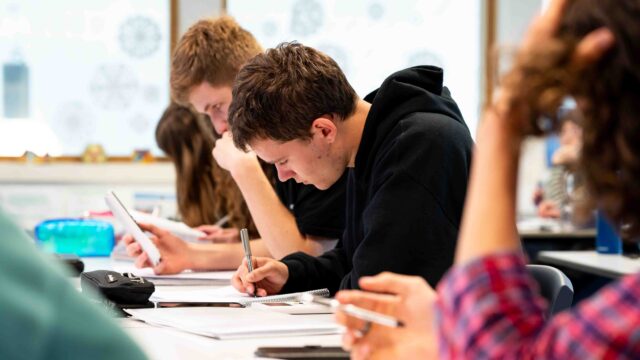 Students sit at a classroom table and focus on their work with pen in hand and notebooks open.