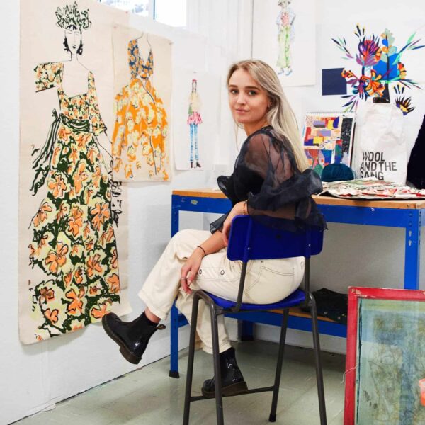 A student, Matilda, sits on a chair looking back at the camera over her shoulder. She wears a black top, white trousers and black boots. There is art work surrounding her.
