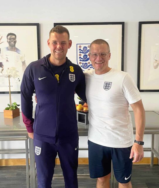 Two people in football kit smile and put their arms around each other.