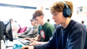 A student sits at a computer with headphones on. He has red hair and a blue jumper.