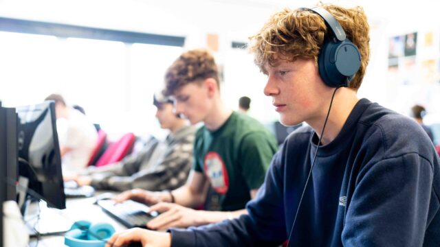 A student sits at a computer with headphones on. He has red hair and a blue jumper.