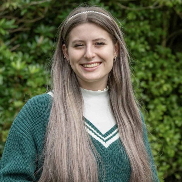 Student, Milarna, stands outside with her hands clasped in front of her. She has long hair and a green jumper and smiles at the camera.
