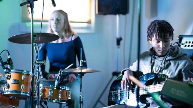 Two students are in a music studio playing instruments. One is playing the drums while the other is playing the guitar.