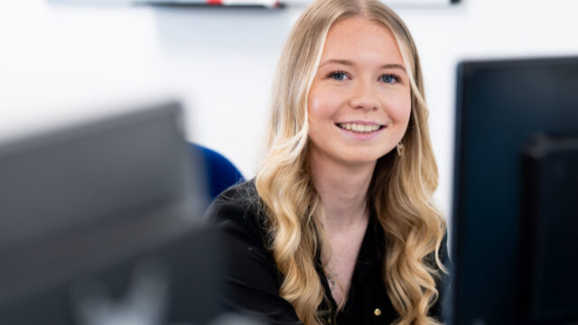 A picture of Olivia at her desk in front of a computer looking at the camera and smiling.