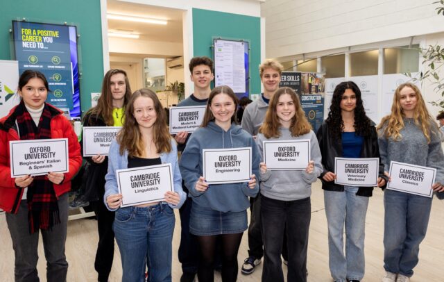 A group of students stand holding bits of paper that says which University they are attending and what they are studying.