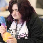 A student is practicing creating tattoos on an orange. The student has purple hair and is wearing a black hoodie.