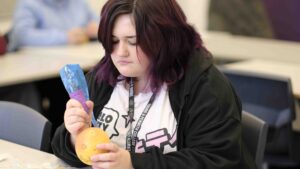 A student is practicing creating tattoos on an orange. The student has purple hair and is wearing a black hoodie.