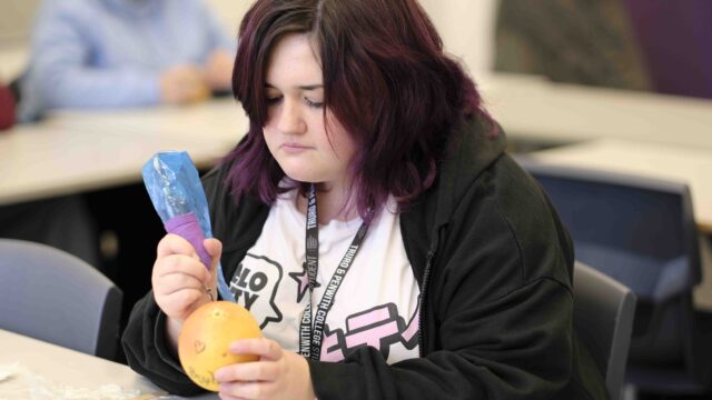 A student is practicing creating tattoos on an orange. The student has purple hair and is wearing a black hoodie.