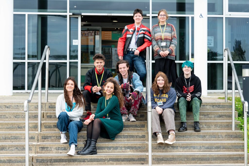 A group of students sit on the steps of the Lynher building at Truro College. The students all have a unique style and are wearing lanyards. They all smile at the camera.
