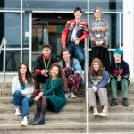 A group of students sit on the steps of the Lynher building at Truro College. The students all have a unique style and are wearing lanyards. They all smile at the camera.