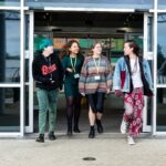 A group of students are exiting a building on Truro campus, chatting and laughing. They are all wearing bright and unique clothing with college lanyards.