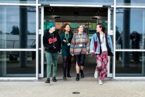 A group of students are exiting a building on Truro campus, chatting and laughing. They are all wearing bright and unique clothing with college lanyards.