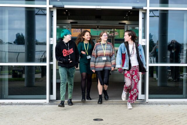 A group of students are exiting a building on Truro campus, chatting and laughing. They are all wearing bright and unique clothing with college lanyards.