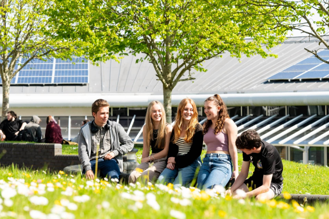 A group of students sit outside at Penwith College leaning against a wall on a sunny day. There is bright green grass and daisies in the foreground. The students laugh and smile.