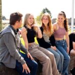 Five students sit on a low wall chatting and smiling. It's a sunny day at Penwith College and the grass is very green.