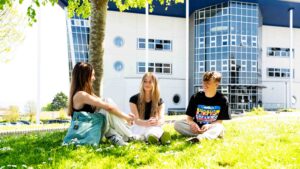 Three students sit on the grass outside a building at Penwith College. The sun is shining and there are daisies surrounding them.