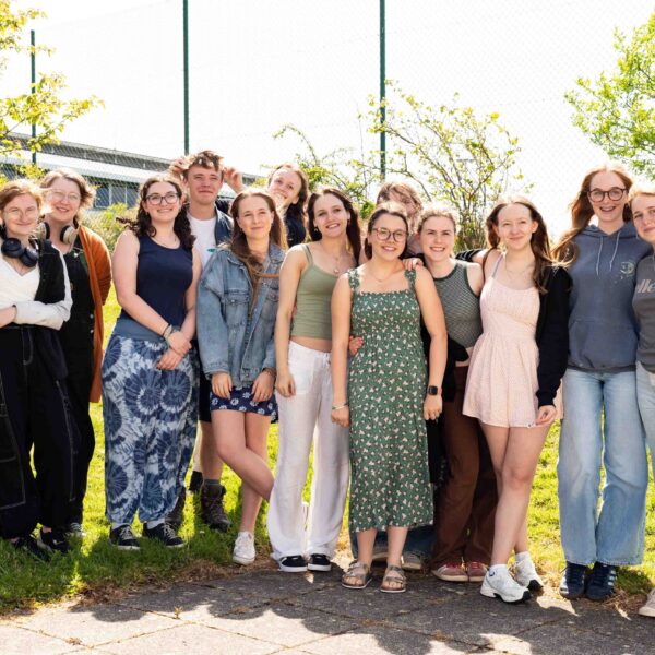A large group of students stand in a line and smile at the camera. Some students have their arms around each other and some are resting their heads on each others shoulders. It is a sunny day and there are trees in the background.