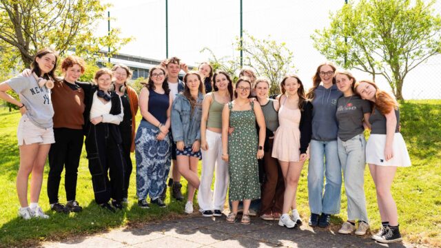 A large group of students stand in a line and smile at the camera. Some students have their arms around each other and some are resting their heads on each others shoulders. It is a sunny day and there are trees in the background.