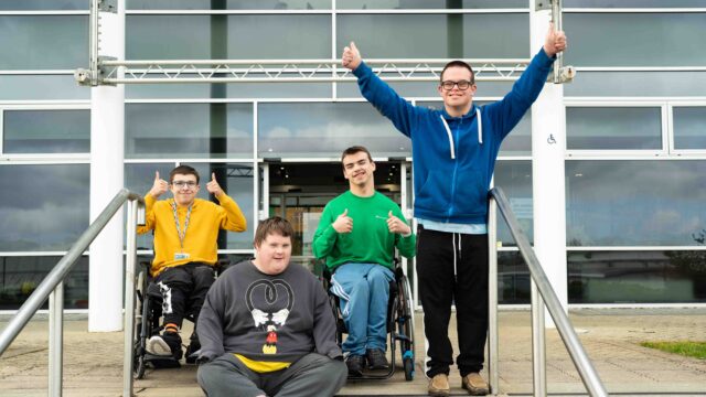Four progression pathways students are at the steps of the Lynher building. They put their thumbs up to the camera. Two of the students are in wheelchairs.