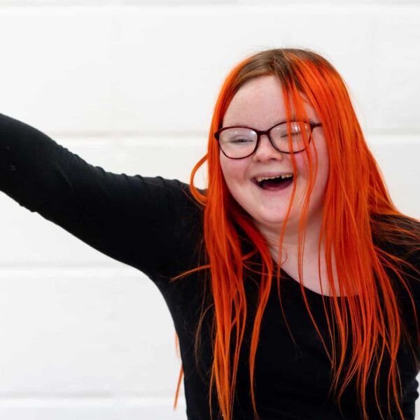 A student with long red hair and glasses has their hand in the air and are laughing.