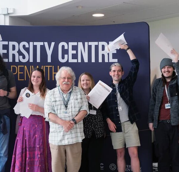 A group of University students hold their results up in the air and their tutor stands in front of them.