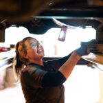 A female automotive student looks at the underneath of a car. She has blue gloves on and glasses and is smiling.