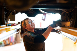 A female automotive student looks at the underneath of a car. She has blue gloves on and glasses and is smiling.