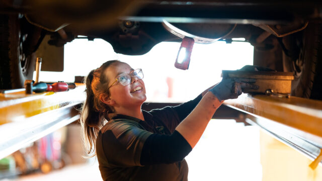 A female automotive student looks at the underneath of a car. She has blue gloves on and glasses and is smiling.