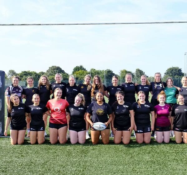 The female rugby team pose underneath a goal post. The front line kneel and the line behind are standing.