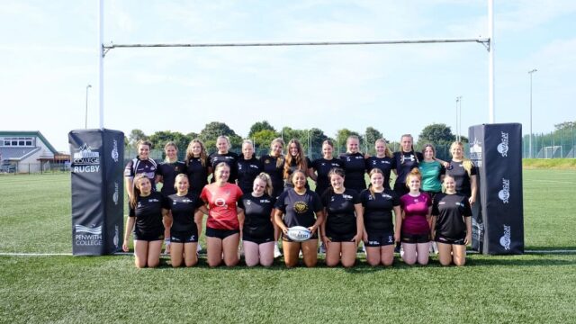 The female rugby team pose underneath a goal post. The front line kneel and the line behind are standing.