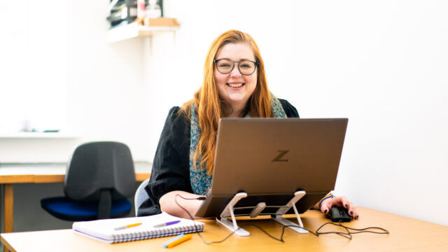 A yound woman with red hair sits at a desk with a laptop in front of her. She looks over the top and smiles.