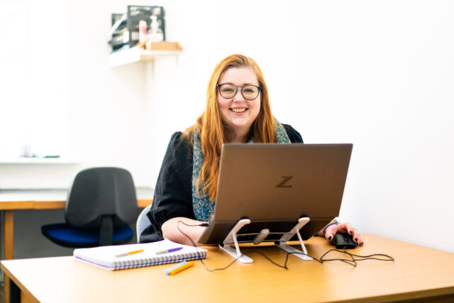 A yound woman with red hair sits at a desk with a laptop in front of her. She looks over the top and smiles.