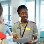 A student nurse is holding paper and looking at fellow students as she is mid-way through speaking. There is branding on her nurse's outfit and she is wearing a lanyard.