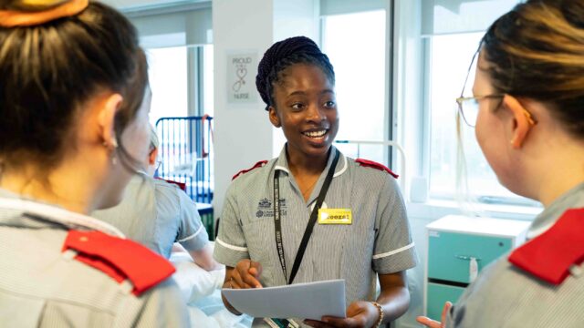 A student nurse is holding paper and looking at fellow students as she is mid-way through speaking. There is branding on her nurse's outfit and she is wearing a lanyard.