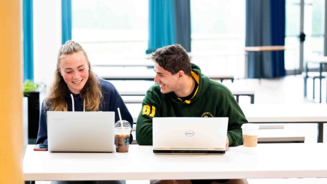 Two students sit at a large table with laptops each. They both have shop bought drinks. They are smiling.