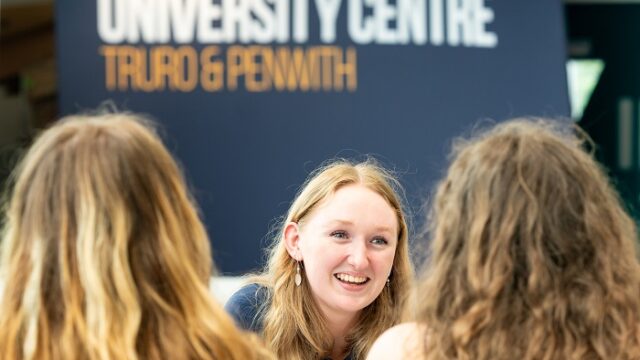 A close up image of a student with a University Centre banner behind her. The student looks at two other students who have their backs to the camera.