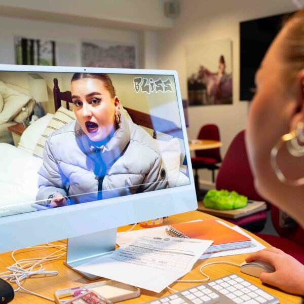 The camera looks over a student's shoulder at a computer screen playing a video. The video shows a person talking wearing a coat. There are notebooks all over the computer desk.
