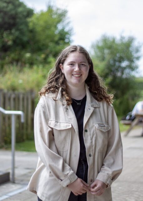Ellie is stood outside at the college with a picnic bench in the background.