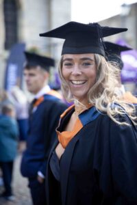 A female graduate turns her head towards the camera and smiles. She is in a cap and gown and the wind is blowing her hair.