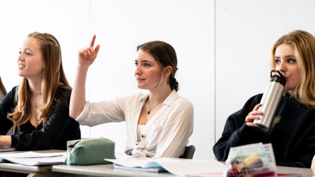 Students sat at classroom tables looking towards the front. The student in the centre has her hand up in the air.