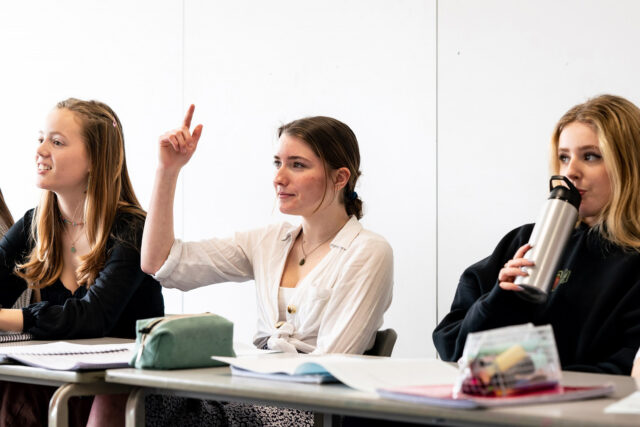 Students sat at classroom tables looking towards the front. The student in the centre has her hand up in the air.