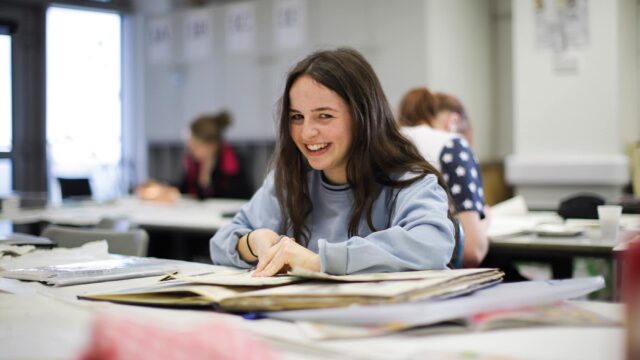 A student works in a sketchbook and looks up at the camera and smiles.
