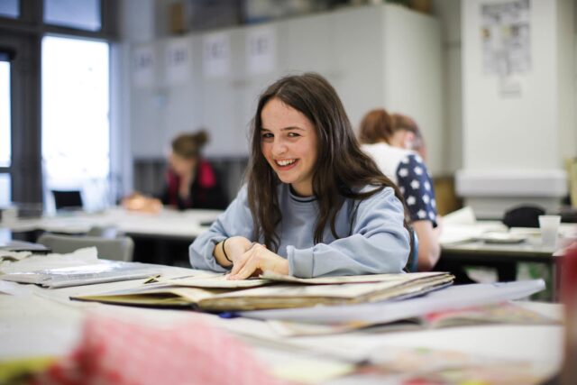 A student works in a sketchbook and looks up at the camera and smiles.