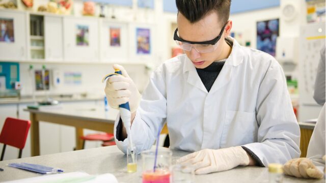 A student in a lab coat with beakers on a table in front of them.
