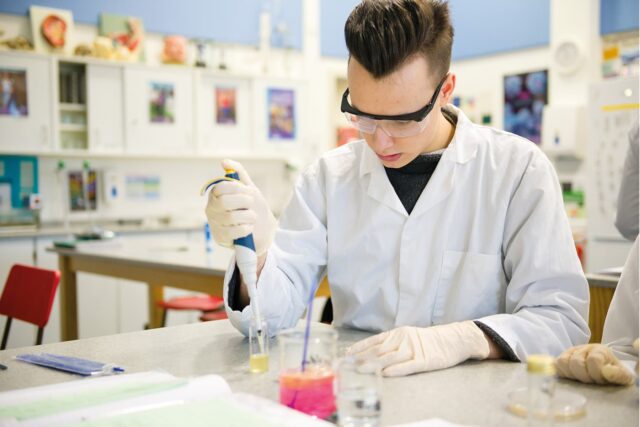 A student in a lab coat with beakers on a table in front of them.
