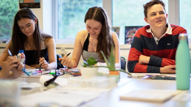 Three students sat at classroom tables with notepads, pens and water bottles.