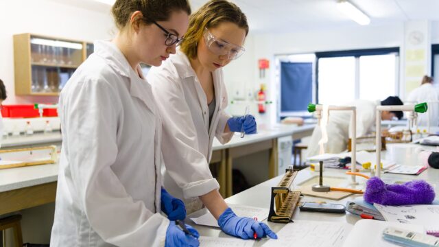 Two students in lab coats are looking at paperwork in a laboratory.