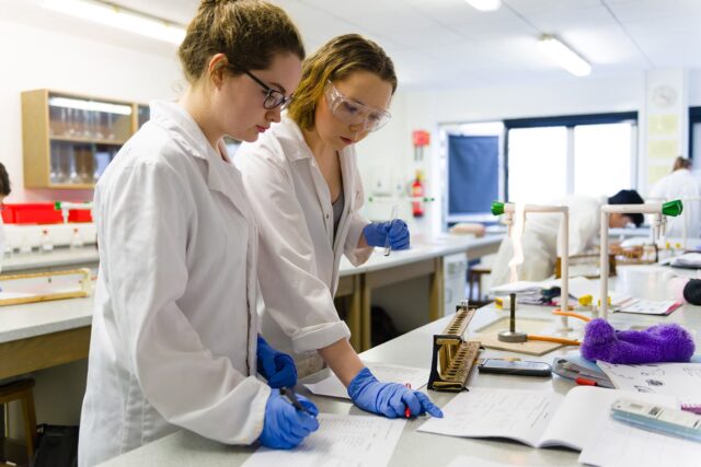 Two students in lab coats are looking at paperwork in a laboratory.