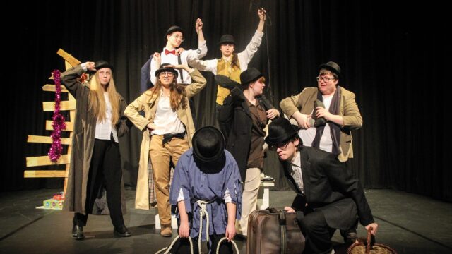 A group of performance students in various costumes on a dark stage.