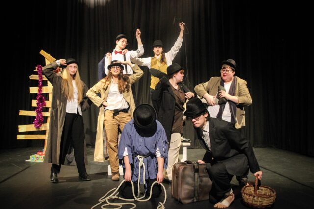 A group of performance students in various costumes on a dark stage.
